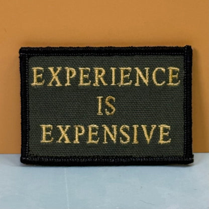Front view of Experience Is Expensive patch in olive green with yellow lettering