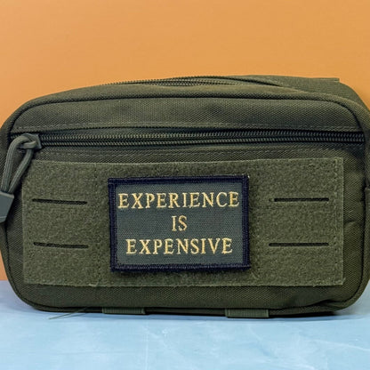 Experience Is Expensive patch in olive green with yellow lettering, mounted on an olive utility pouch