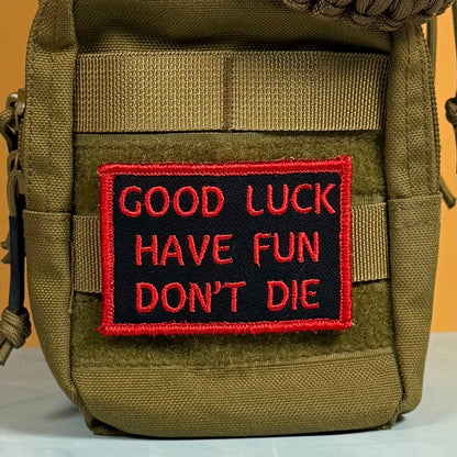 Good Luck, Have Fun, Don't Die patch in black with red lettering and border, mounted on an olive backpack
