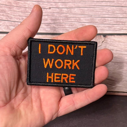 I Don't Work Here patch in black with orange lettering, held in a hand