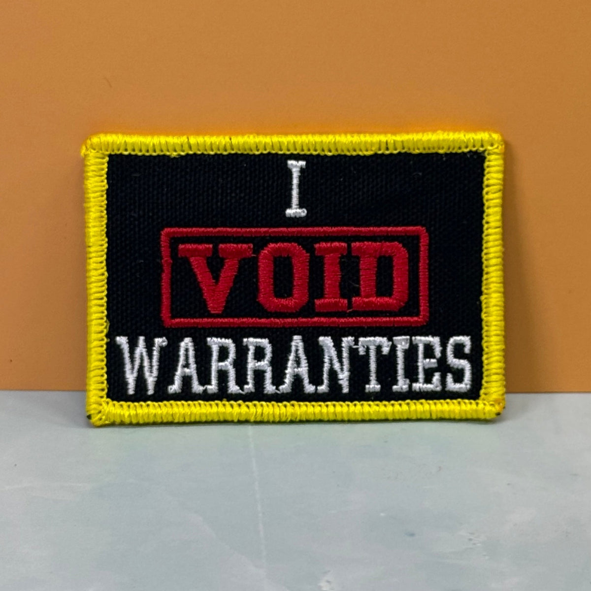 Front view of I Void Warranties patch in black with a red VOID stamp, white lettering and yellow border