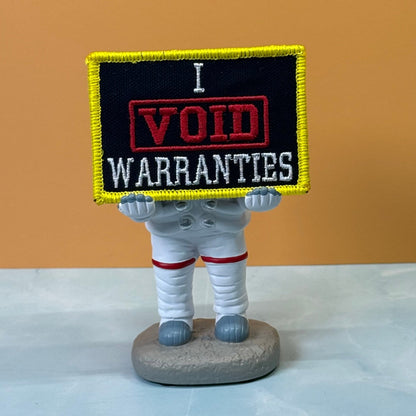 I Void Warranties patch in black with a red VOID stamp, white lettering and yellow border, propped on a small astronaut figurine