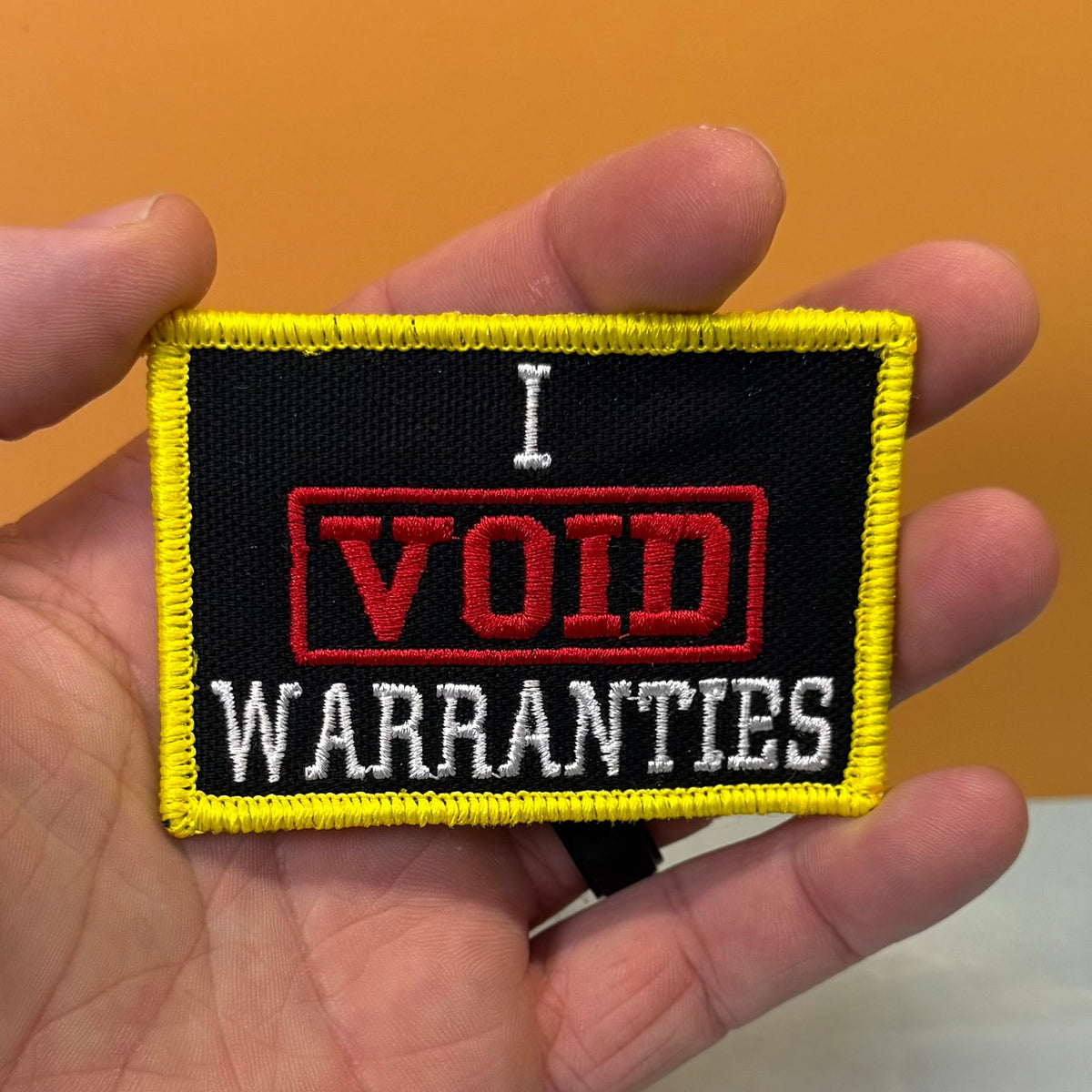 I Void Warranties patch in black with a red VOID stamp, white lettering and yellow border, held in a hand