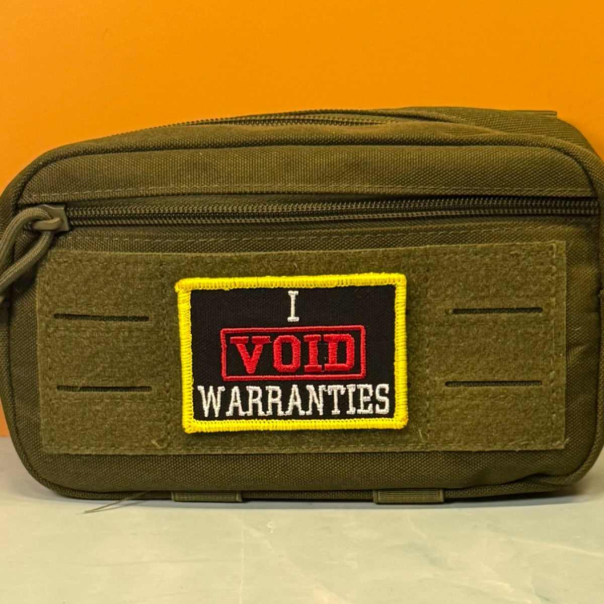 I Void Warranties patch in black with a red VOID stamp, white lettering and yellow border, mounted on an olive utility pouch