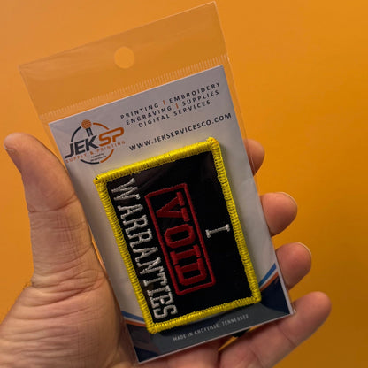I Void Warranties patch in black with a red VOID stamp, white lettering and yellow border, retail package held in a hand