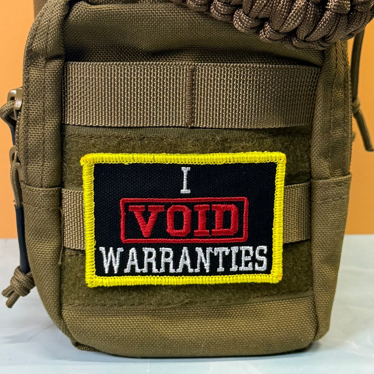 I Void Warranties patch in black with a red VOID stamp, white lettering and yellow border, mounted on an olive backpack