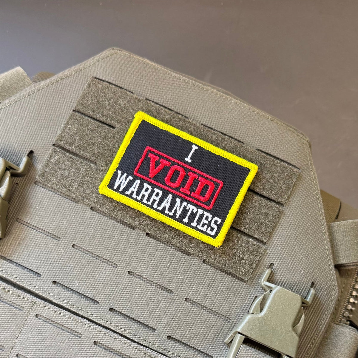 I Void Warranties patch in black with a red VOID stamp, white lettering and yellow border, mounted on a tan plate carrier