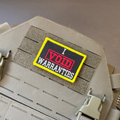 I Void Warranties patch in black with a red VOID stamp, white lettering and yellow border, mounted on a tan plate carrier