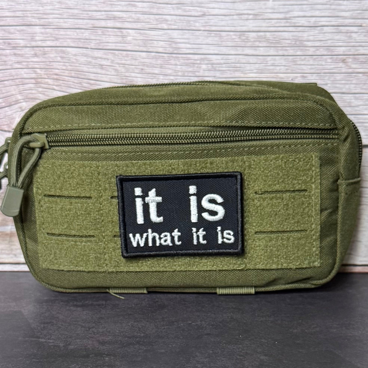 It Is What It Is patch in black with white lowercase lettering, mounted on an olive utility pouch