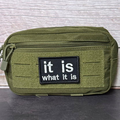 It Is What It Is patch in black with white lowercase lettering, mounted on an olive utility pouch