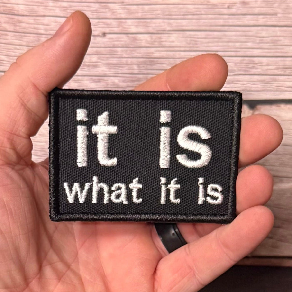It Is What It Is patch in black with white lowercase lettering, held in a hand