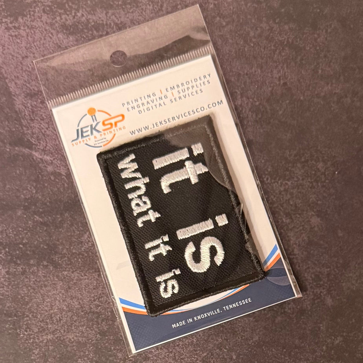 It Is What It Is patch in black with white lowercase lettering, in clear retail packaging