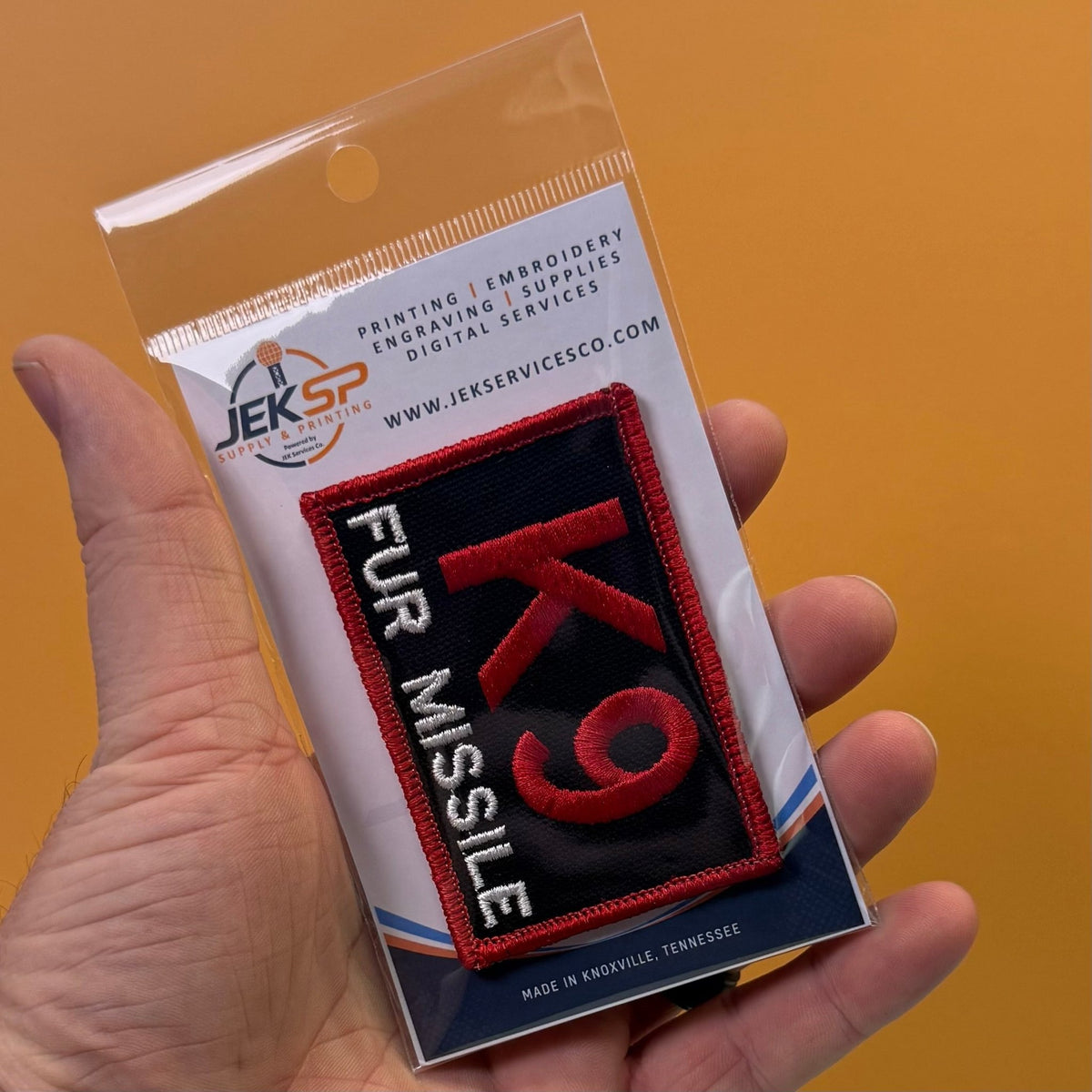 K-9 Fur Missile patch in black with red K9 and border and white FUR MISSILE lettering, retail package held in a hand