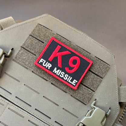 K-9 Fur Missile patch in black with red K9 and border and white FUR MISSILE lettering, mounted on a tan plate carrier