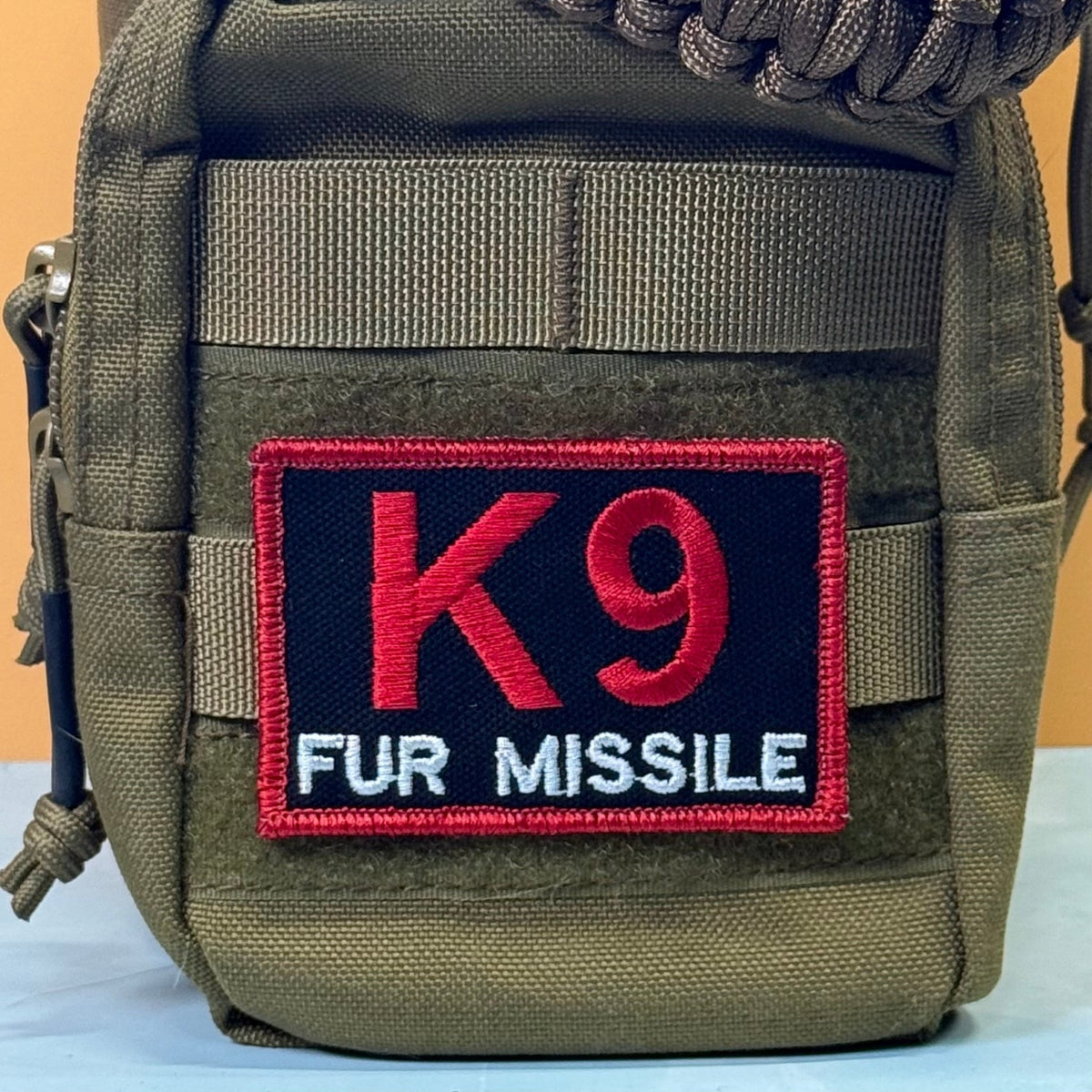 K-9 Fur Missile patch in black with red K9 and border and white FUR MISSILE lettering, mounted on an olive backpack