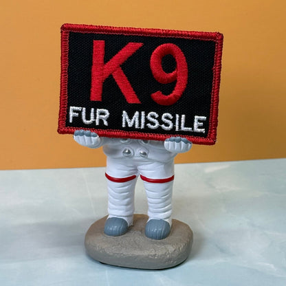 K-9 Fur Missile patch in black with red K9 and border and white FUR MISSILE lettering, propped on a small astronaut figurine