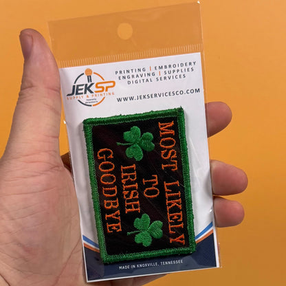 Most Likely To Irish Goodbye patch in black with gold lettering, green shamrocks and a green border, retail package held in a hand