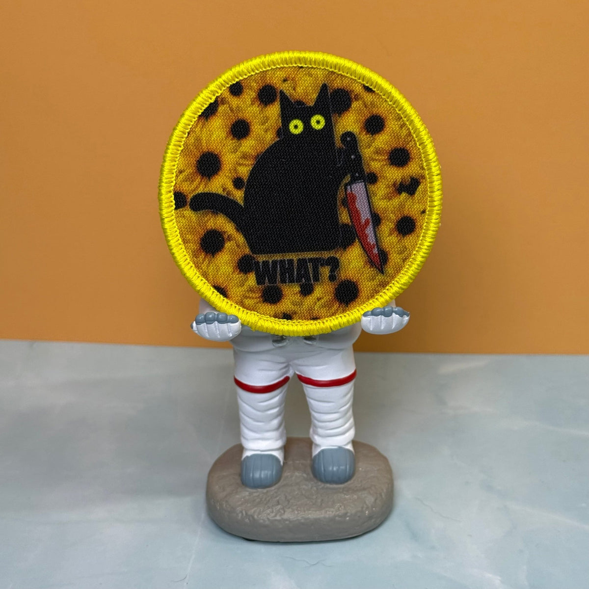 Murder Cat & Sunflowers patch in yellow with a black cat, sunflowers and a red knife, propped on a small astronaut figurine