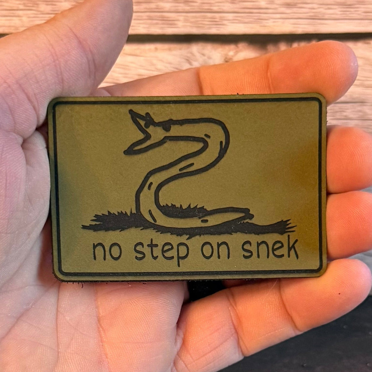 No Step On Snek patch in olive green with a black snake drawing, held in a hand