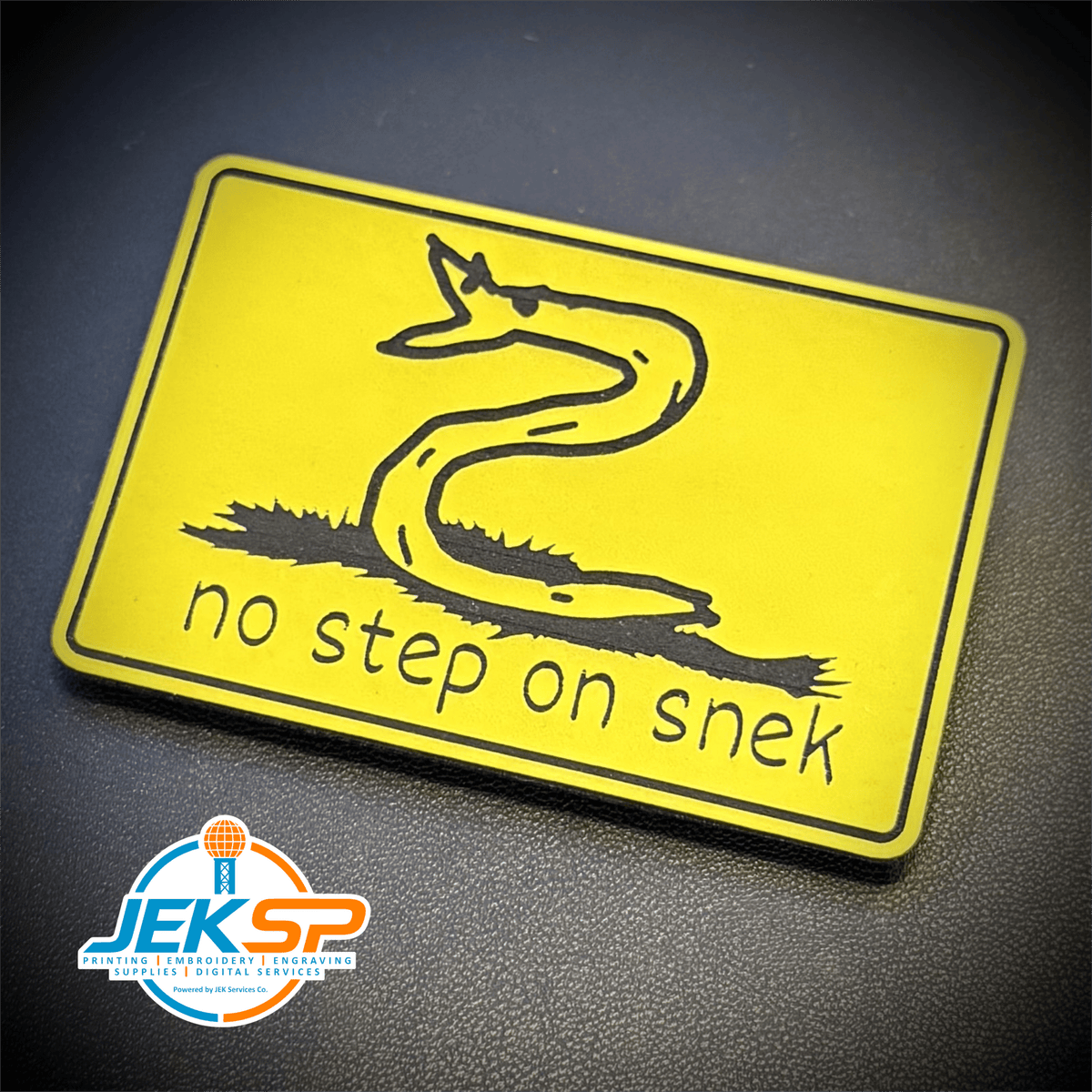 Front view of No Step On Snek patch in yellow with a black snake drawing and lowercase lettering