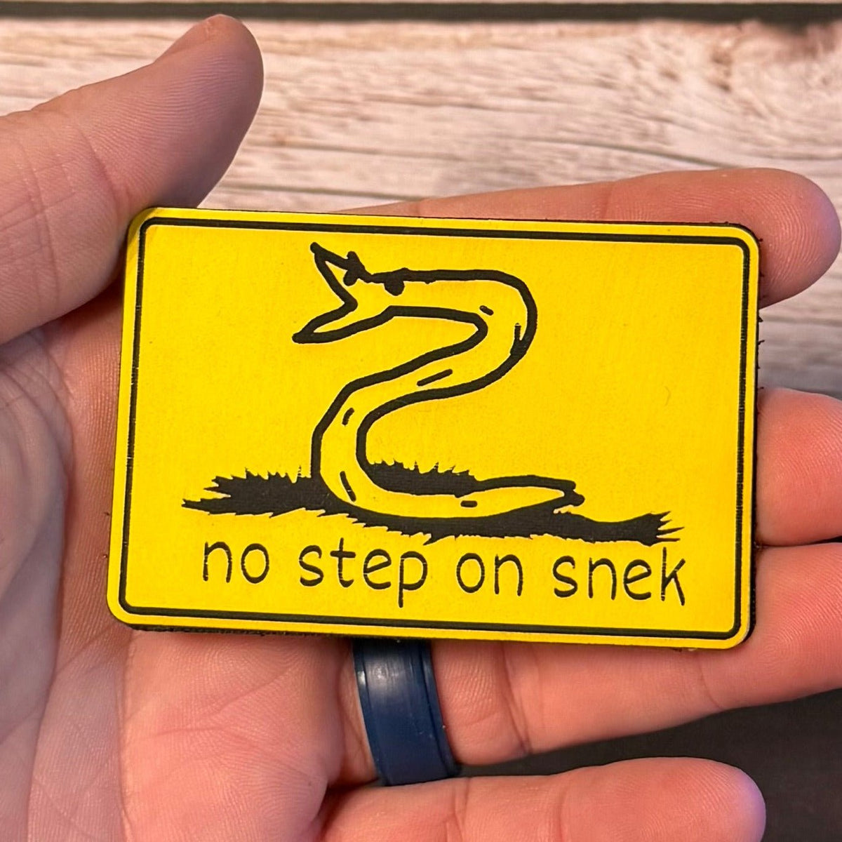 No Step On Snek patch in yellow with a black snake drawing, held in a hand