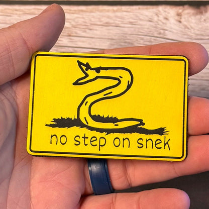 No Step On Snek patch in yellow with a black snake drawing, held in a hand