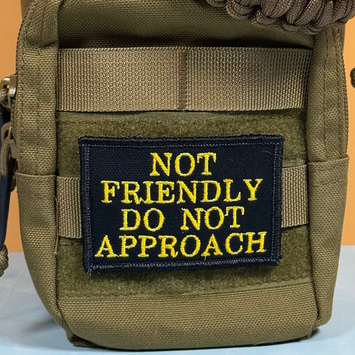 Not Friendly, Do Not Approach patch in black with yellow lettering, mounted on an olive backpack