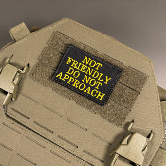 Not Friendly, Do Not Approach patch in black with yellow lettering, mounted on a tan plate carrier
