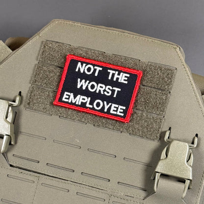 Not The Worst Employee patch in black with white lettering and a red border, mounted on a tan plate carrier