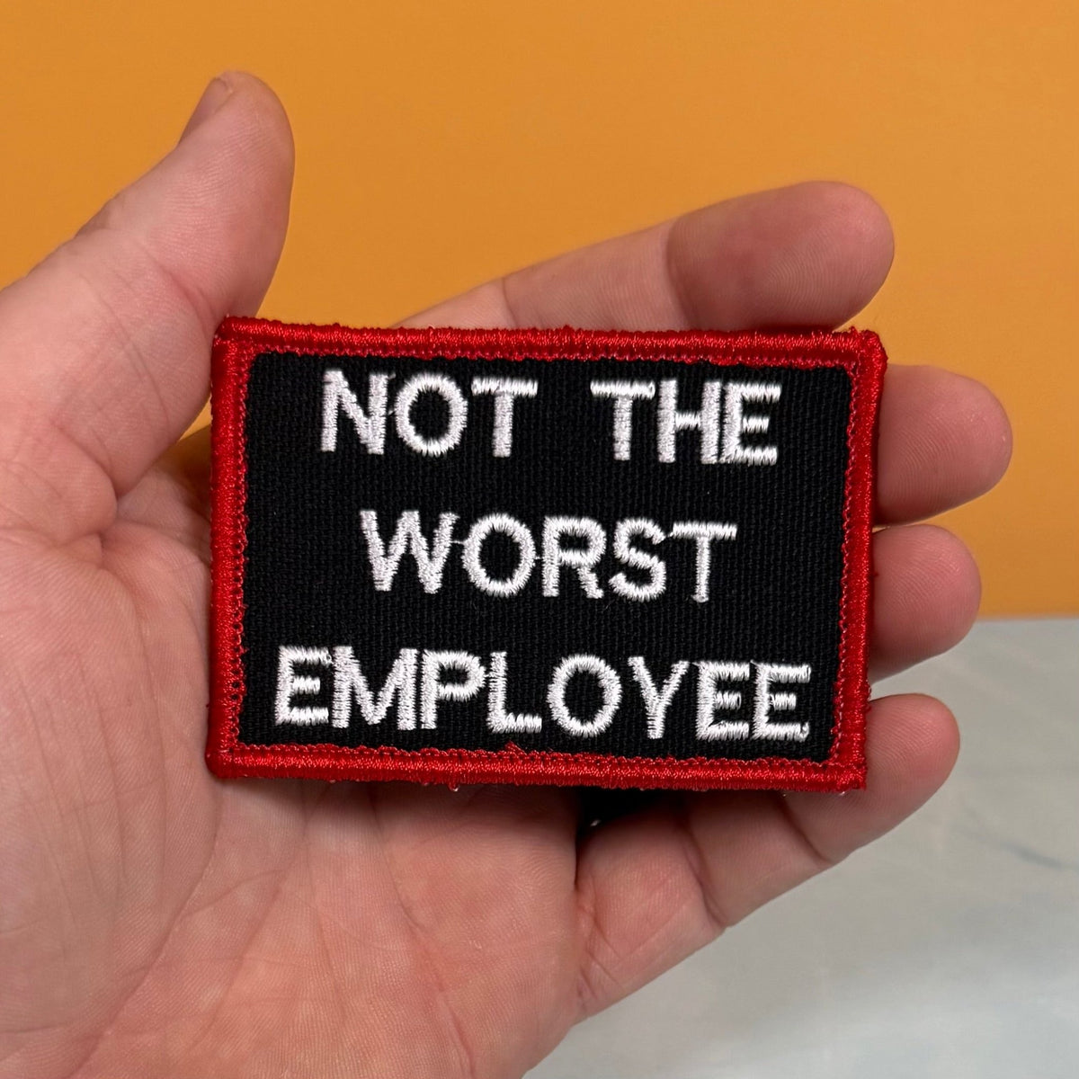 Not The Worst Employee patch in black with white lettering and a red border, held in a hand