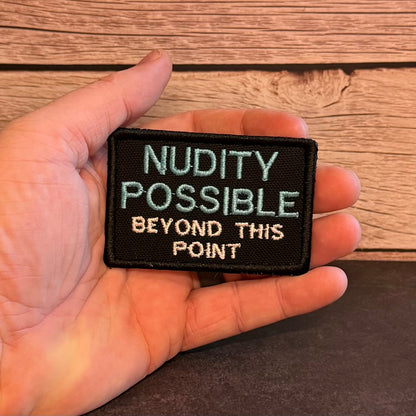 Nude Beach Warning patch in black with blue NUDITY POSSIBLE and white lettering, held in a hand
