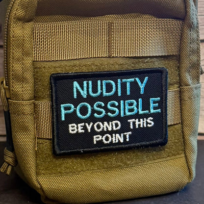 Nude Beach Warning patch in black with blue NUDITY POSSIBLE and white lettering, mounted on an olive backpack