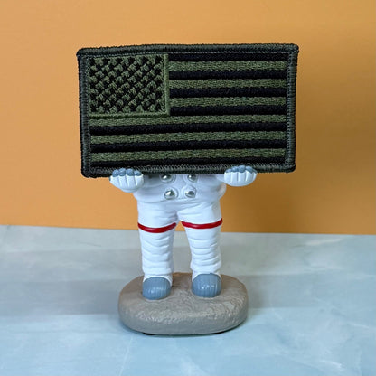 Olive OD Green Tactical American Flag patch in olive green and black stars and stripes, propped on a small astronaut figurine
