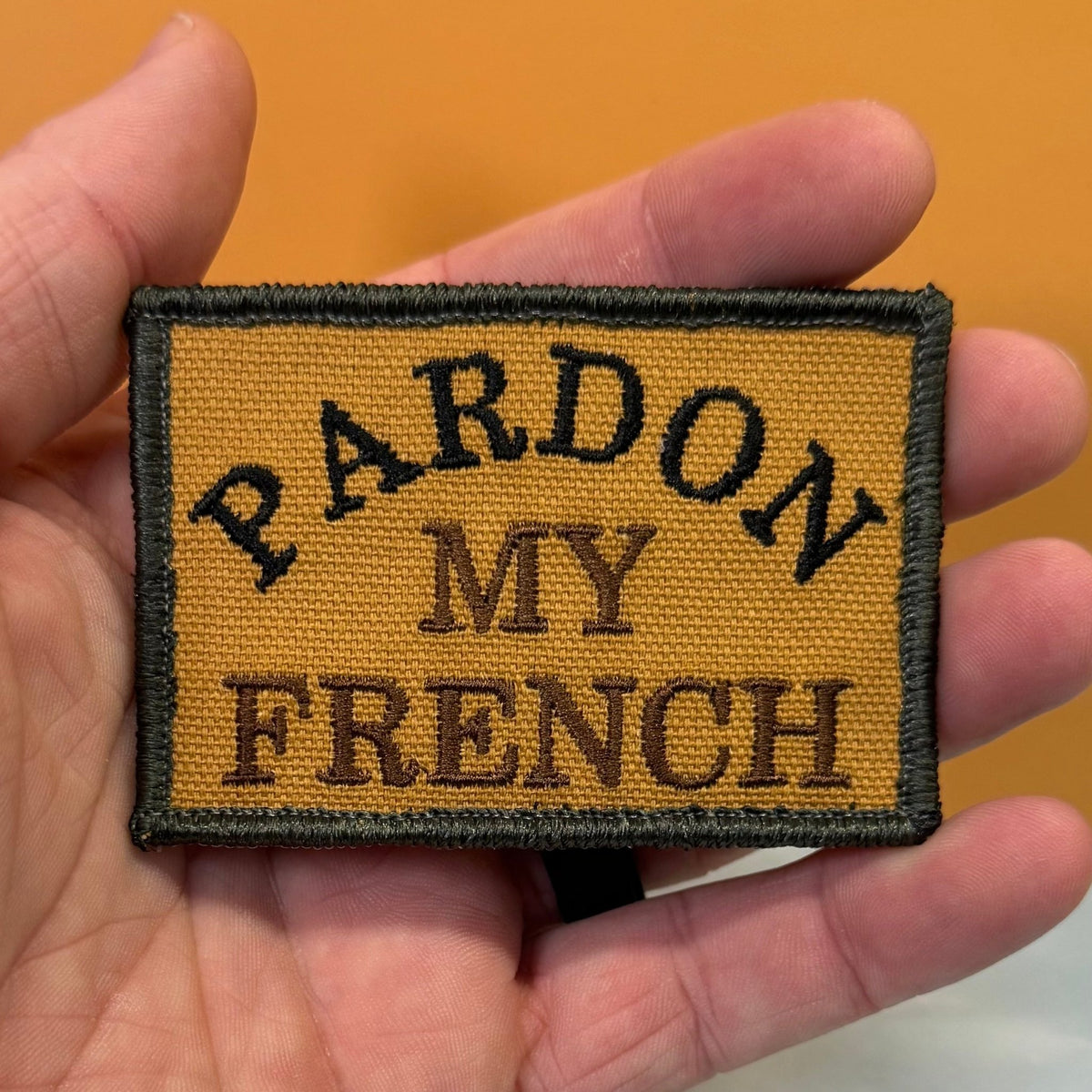 Pardon My French patch in tan with black lettering and border, held in a hand