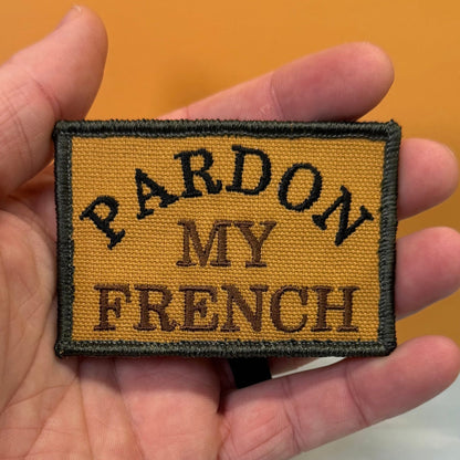 Pardon My French patch in tan with black lettering and border, held in a hand