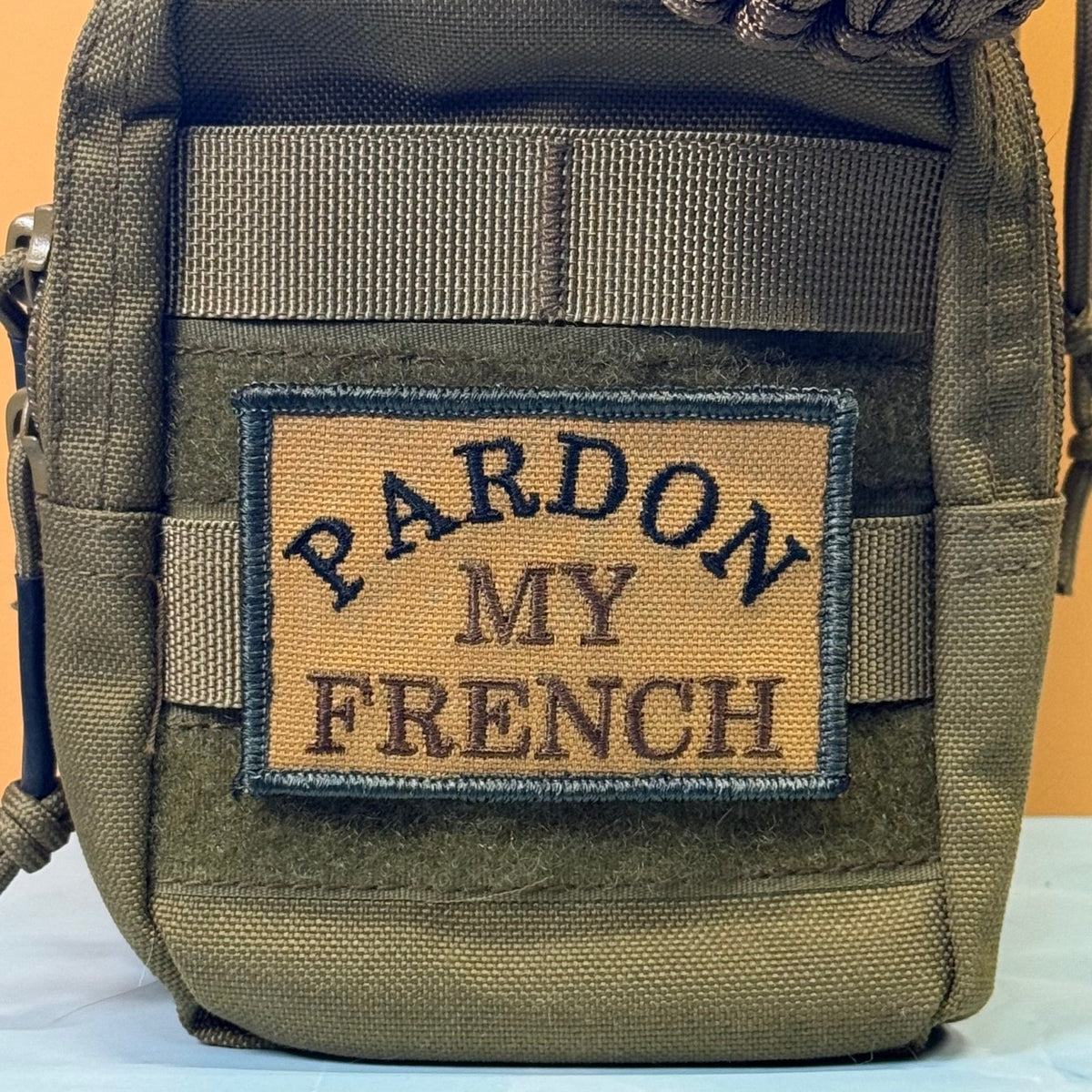Pardon My French patch in tan with black lettering and border, mounted on an olive backpack