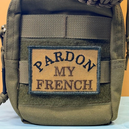 Pardon My French patch in tan with black lettering and border, mounted on an olive backpack