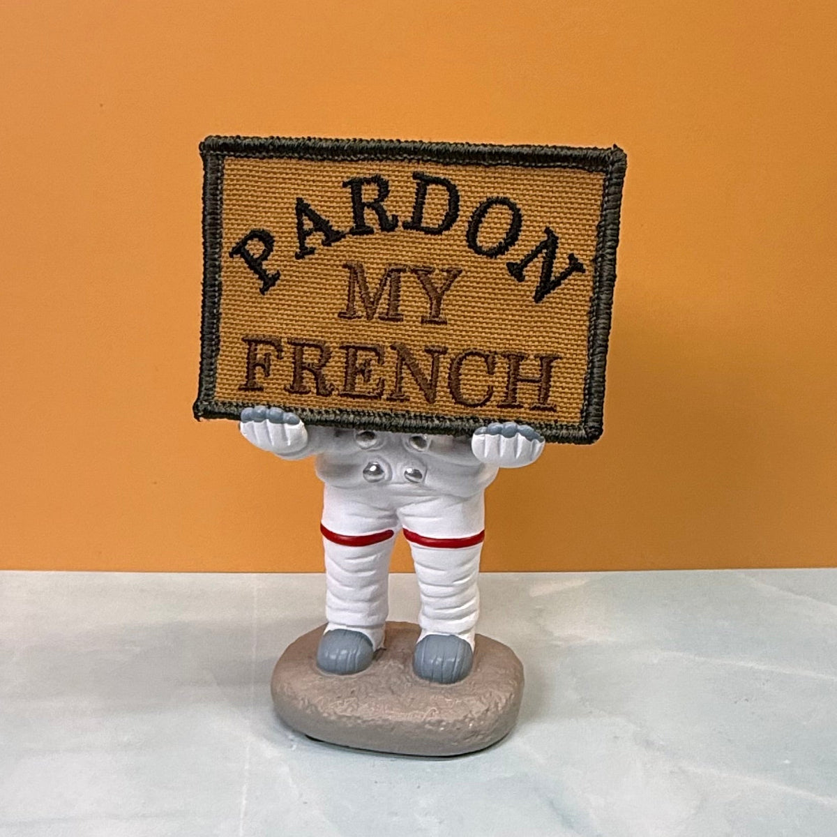 Pardon My French patch in tan with black lettering and border, propped on a small astronaut figurine