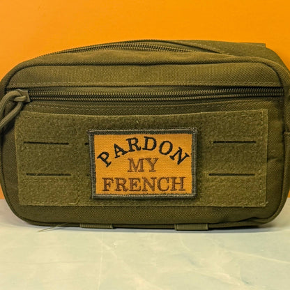 Pardon My French patch in tan with black lettering and border, mounted on an olive utility pouch