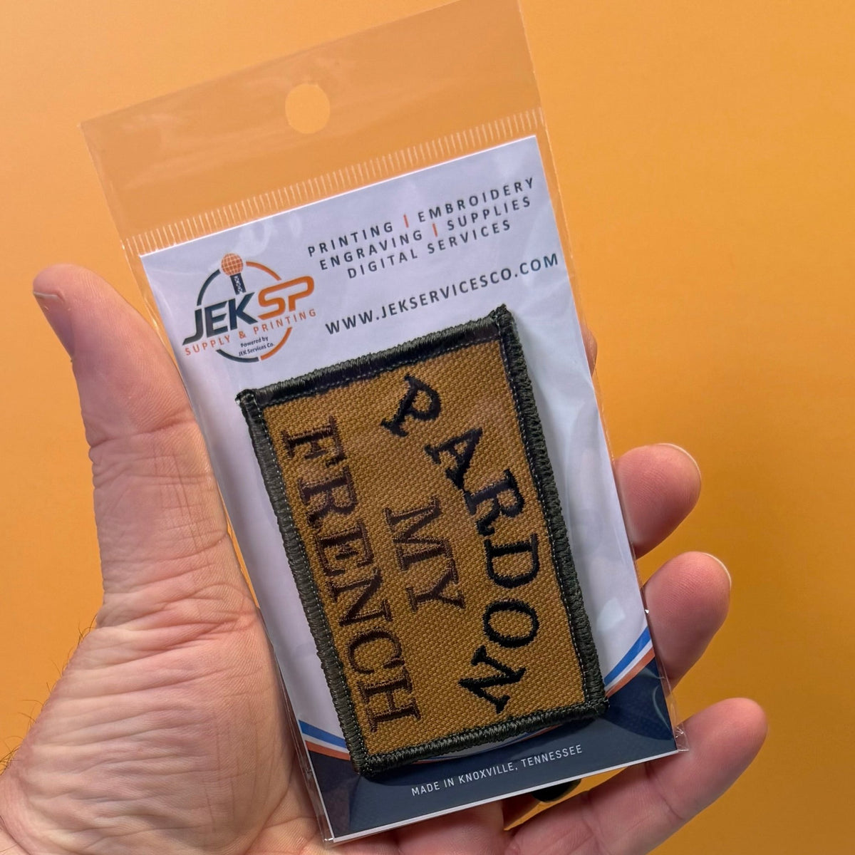 Pardon My French patch in tan with black lettering and border, retail package held in a hand