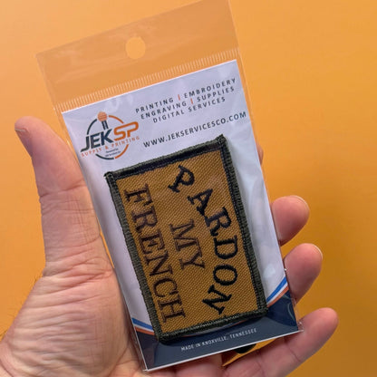 Pardon My French patch in tan with black lettering and border, retail package held in a hand