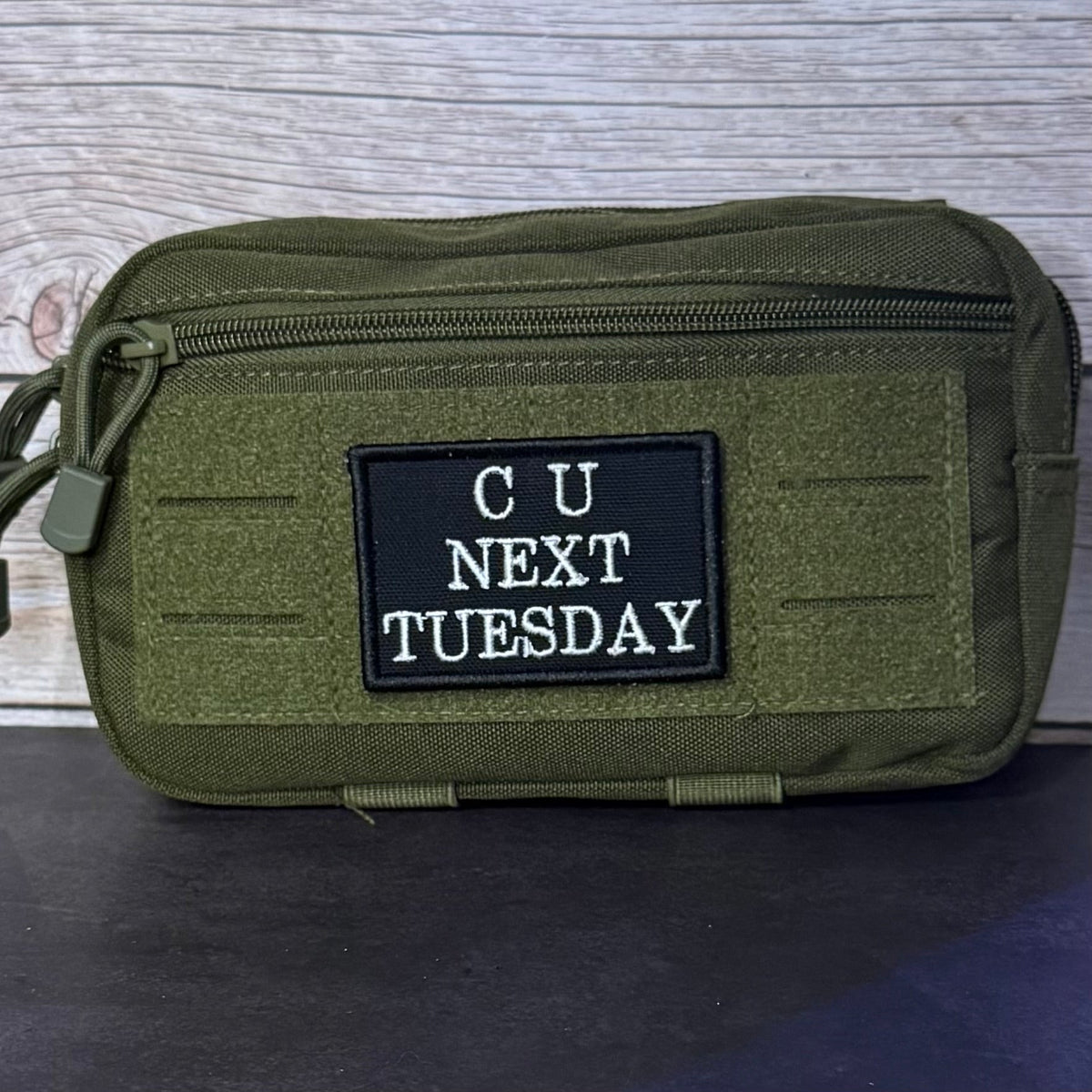 See You Next Tuesday patch in black with white C U NEXT TUESDAY lettering, mounted on an olive utility pouch