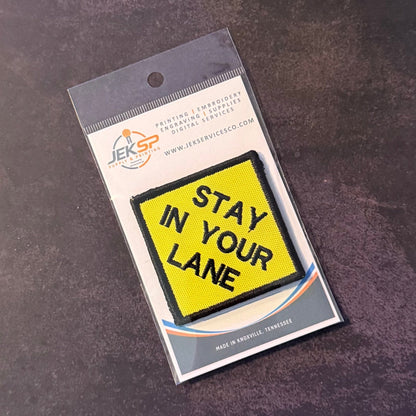 Stay In Your Lane patch in yellow diamond with black lettering and border, in clear retail packaging