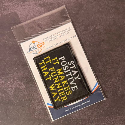 Stay Positive patch in black with white and yellow lettering, in clear retail packaging