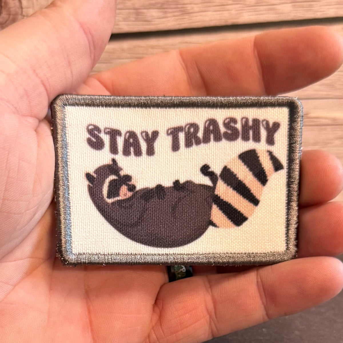 Stay Trashy Raccoon Trash Panda patch in cream with a gray raccoon and dark STAY TRASHY lettering, held in a hand