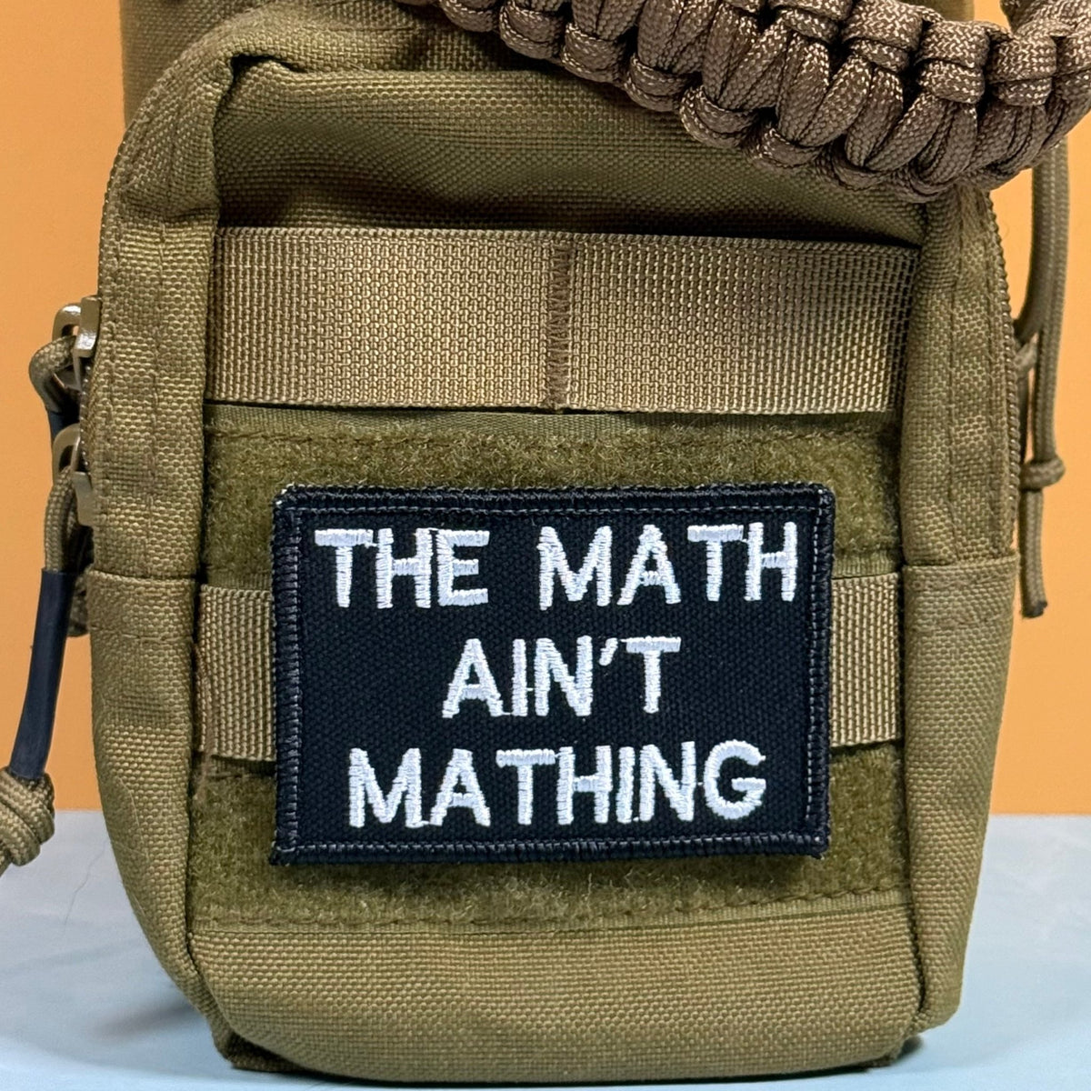 The Math Ain't Mathing patch in black with white lettering, mounted on an olive backpack