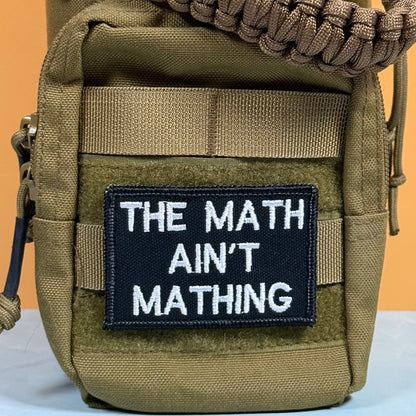The Math Ain't Mathing patch in black with white lettering, mounted on an olive backpack