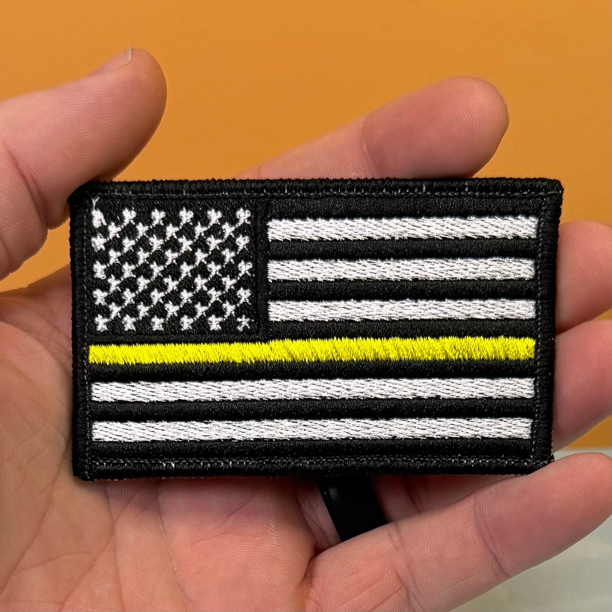 Thin Yellow Line American Flag patch in black and white American flag with one yellow stripe, held in a hand