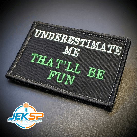 Front view of Underestimate Me, That'll Be Fun patch in black with white and green lettering