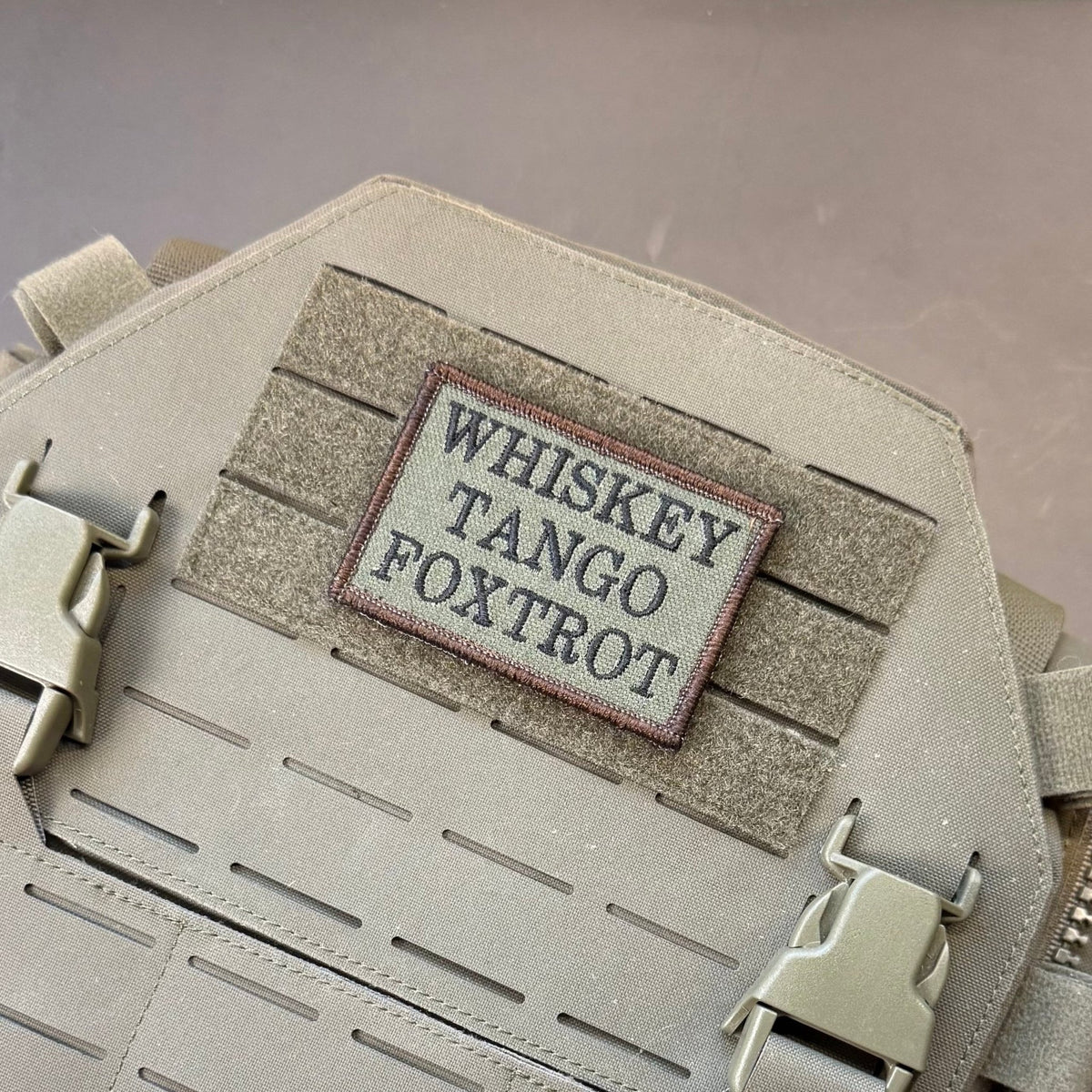 Whiskey Tango Foxtrot WTF patch in olive green with black lettering, mounted on a tan plate carrier